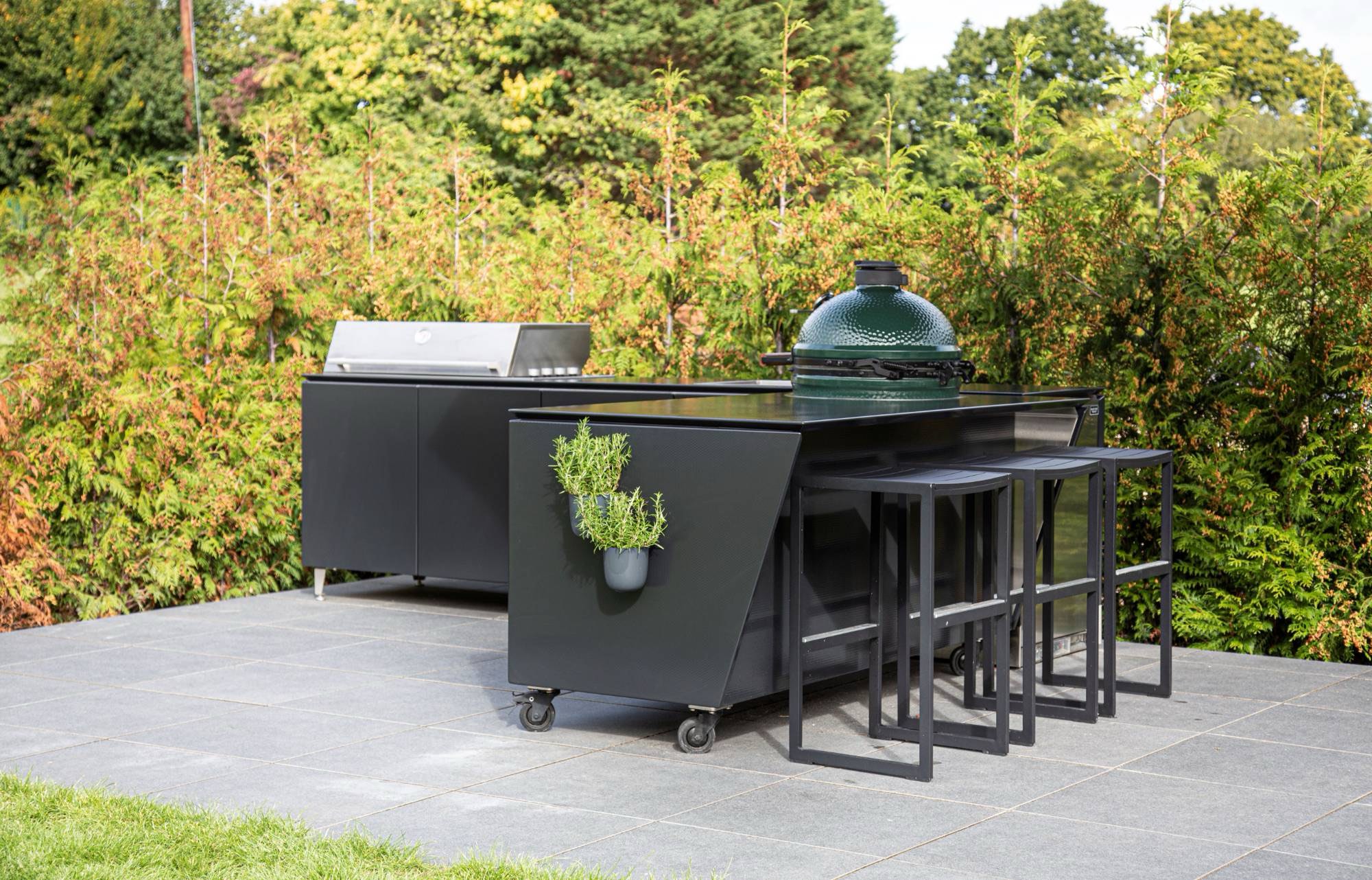 Black mobile outdoor kitchen on locking casters with a ceramic grill, a stainless grill and bar stools on a stone patio