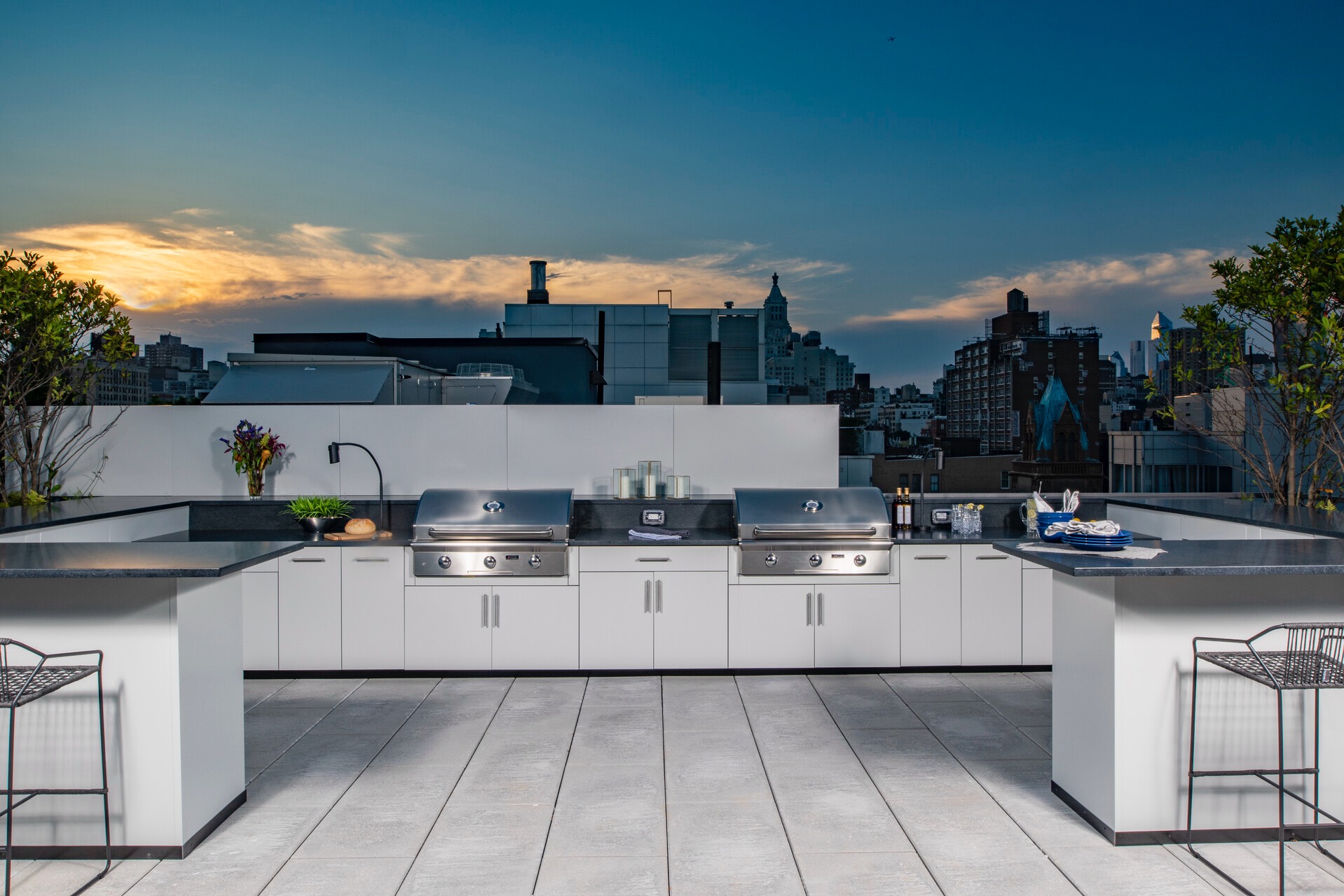 U-shaped white outdoor kitchen with two stainless grills and bar seating on a city rooftop at dusk