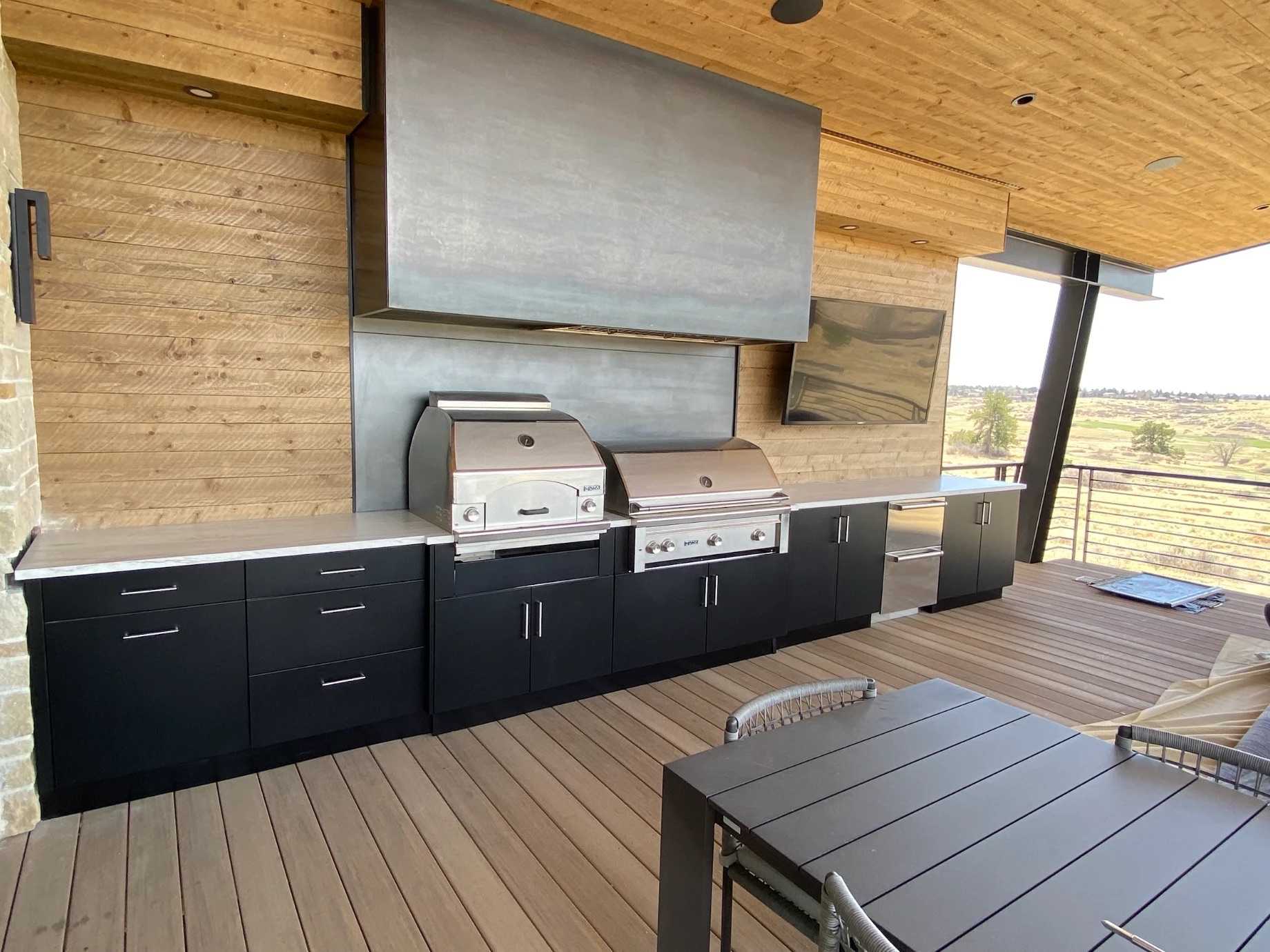 Black outdoor kitchen with a pizza oven and grill on a covered deck overlooking open prairie