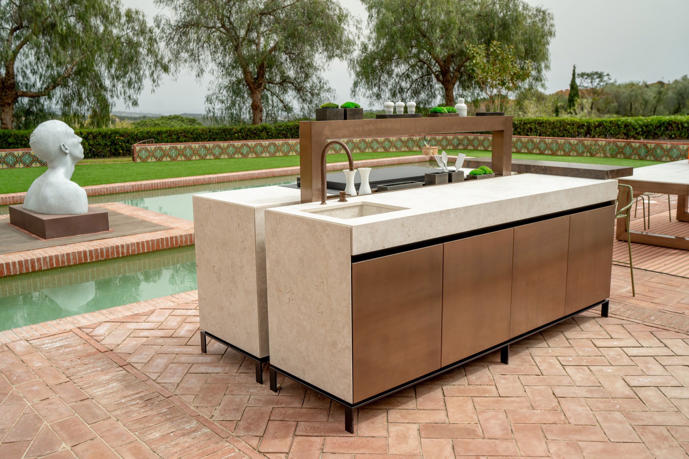 Novara kitchen island with a pale stone top, bronze fronts and a bronze faucet beside a garden pool