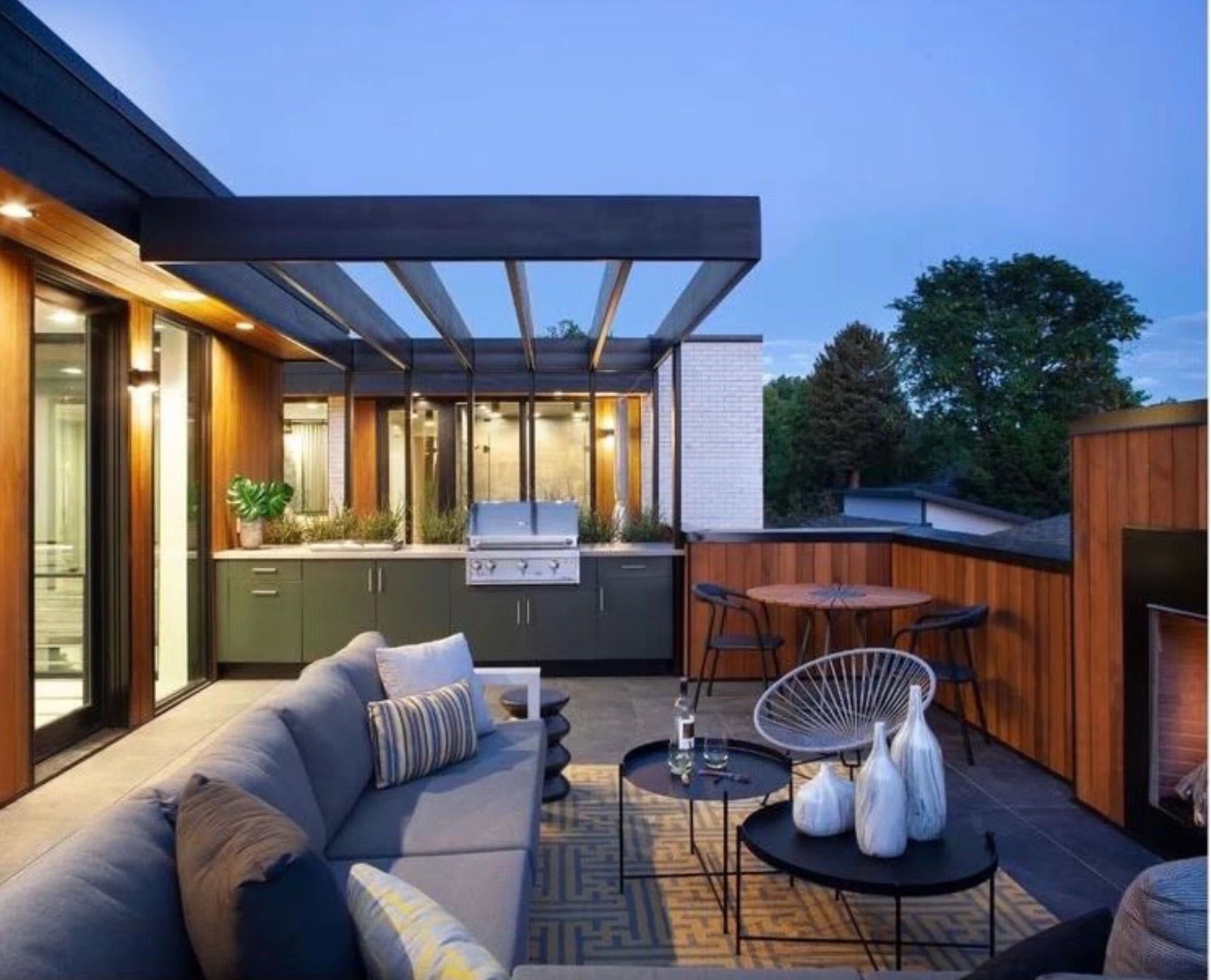 Dark green outdoor kitchen with a built-in grill under a black pergola on a rooftop terrace at dusk